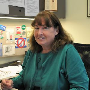 Photo of Marjorie Kruvand, sitting at their desk wearing green sweater